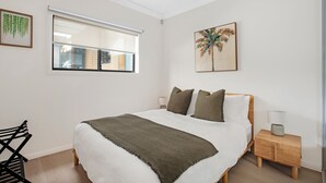 Une chambre à coucher avec un lit, une fenêtre avec des stores, un ventilateur de plafond, une table de chevet et un tableau au mur.