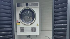 Un espace compact dédié à la buanderie, comprenant deux machines à laver et un sèche-linge, le tout intégré dans une structure grise avec une terrasse en bois à l’extérieur.