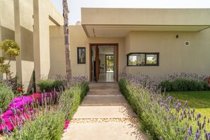 Un bâtiment moderne avec un chemin en béton menant à une porte en bois, le tout entouré de lavande et de fleurs roses.