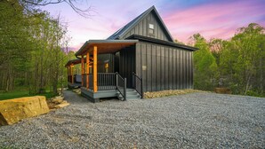 A rustic cabin with a porch, a gravel driveway, and a forested backdrop. - Spiral Loft · Hot Tub · Spa Bath | Lux Sleeps 4 (McArthur)