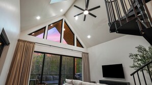A modern living room with a high ceiling, a ceiling fan, a flat-screen TV, a sofa, a dining table, and a staircase. - Spiral Loft · Hot Tub · Spa Bath | Lux Sleeps 4 (McArthur)