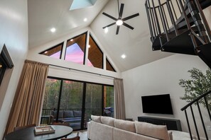 Una sala de estar moderna con techo alto, un ventilador de techo, un televisor de pantalla plana, un sofá, una mesa de comedor y una escalera.