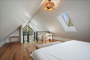 A bedroom with a sloped ceiling, a bed, a chair, a small table, and a window with a view of trees. - Treetop Windows · Hot Tub · Forest Views | Luxe (McArthur)
