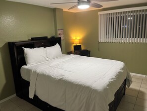 A neatly made bed with white bedding, a ceiling fan, a nightstand with a lamp, and a window with blinds.
