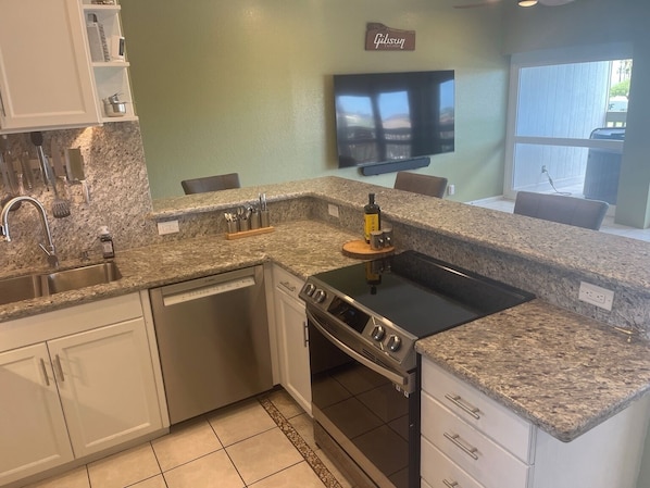 Una cocina con encimeras de granito, lavavajillas, estufa y televisor en la pared.