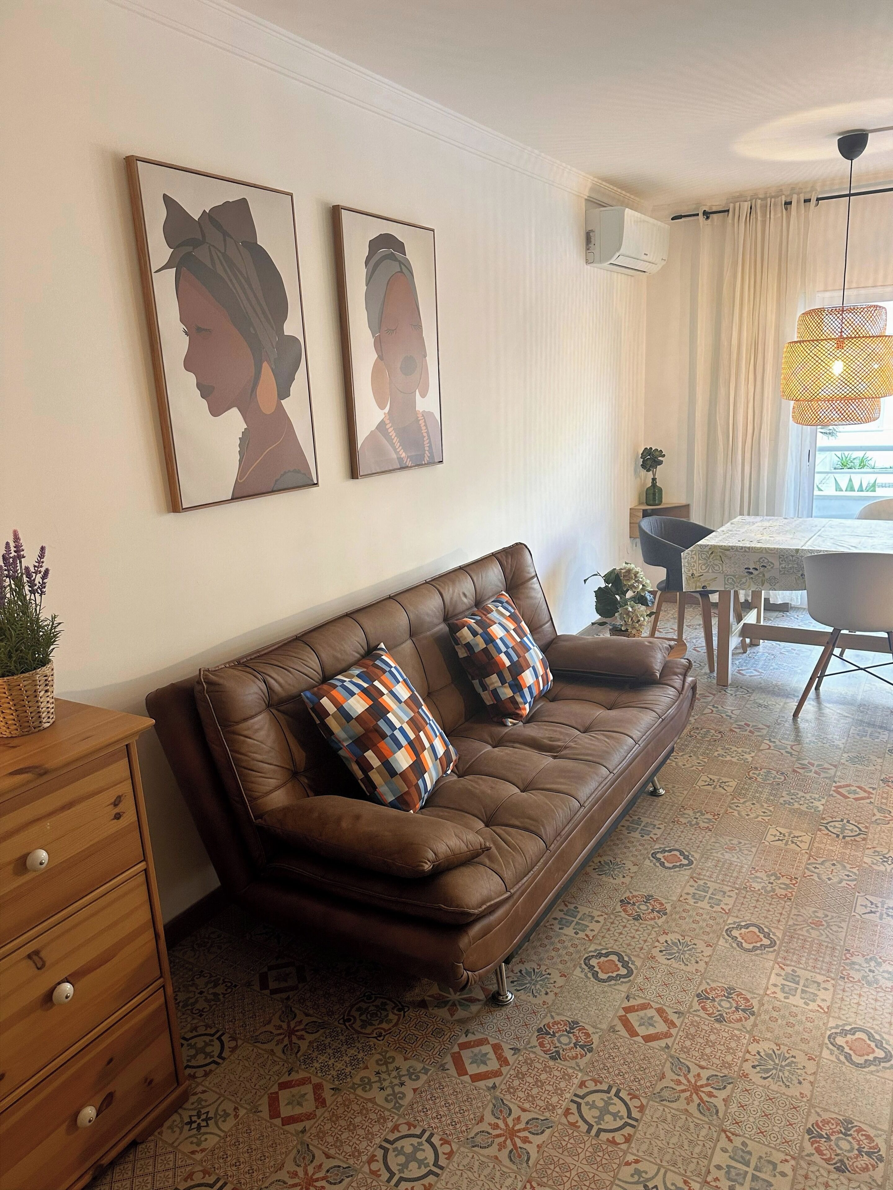 A living room with a brown leather sofa, patterned cushions, a wooden side table, and a dining area with a table and chairs.