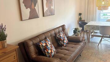 A living room with a brown leather sofa, patterned cushions, a wooden side table, and a dining area with a table and chairs.