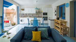 A modern living room with a sofa, a dining area with chairs, a kitchen with white cabinets, and a view through a glass door. - Blue House Relax -Cozy family apartment 3 minutes from the sea (Marina di Ragusa)