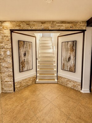 Eine Treppe führt zu einer oberen Etage, begleitet von zwei gerahmten abstrakten Gemälden, mit einer Steinwand und einem gefliesten Boden.