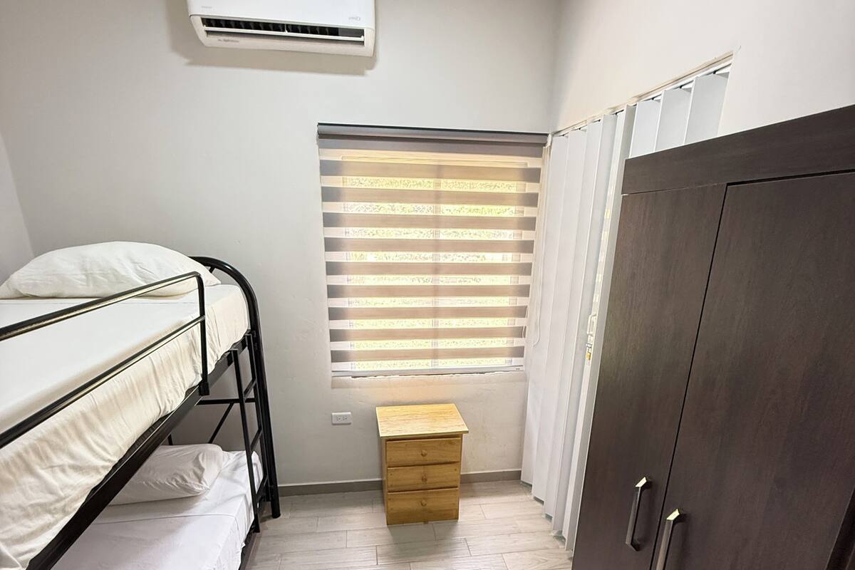 A bedroom with a bunk bed, a small wooden cabinet, a wardrobe, and an air conditioning unit.