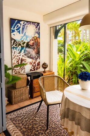 A room with a large poster, wicker furniture, a table with a vase of blue flowers, and a view of greenery outside. - 1-bedroom apartment, beachfront, in a complex with 3 pools and a garden (anse marcel)