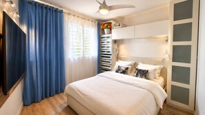 A bedroom with a bed, curtains, a ceiling fan, a window with blinds, and a wall-mounted shelf with flowers. - 1-bedroom apartment, beachfront, in a complex with 3 pools and a garden (anse marcel)