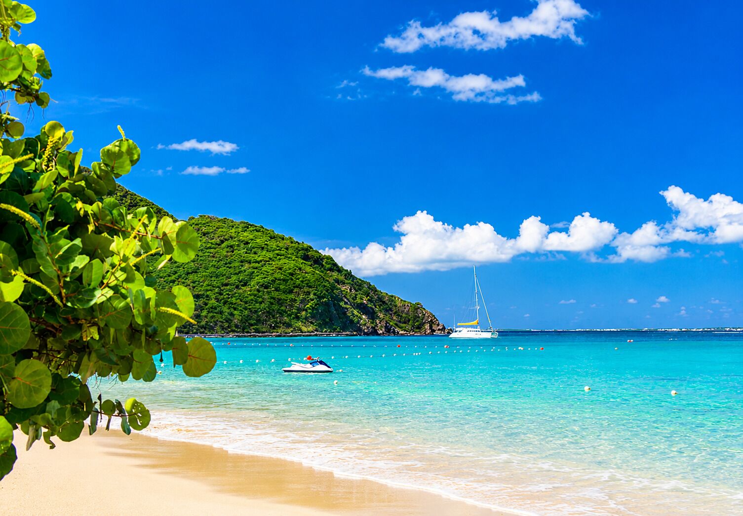 A beach with clear turquoise waters, a sailboat, and a lush green hill in the background.
