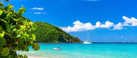 A beach with clear turquoise waters, a sailboat, and a lush green hill in the background.
