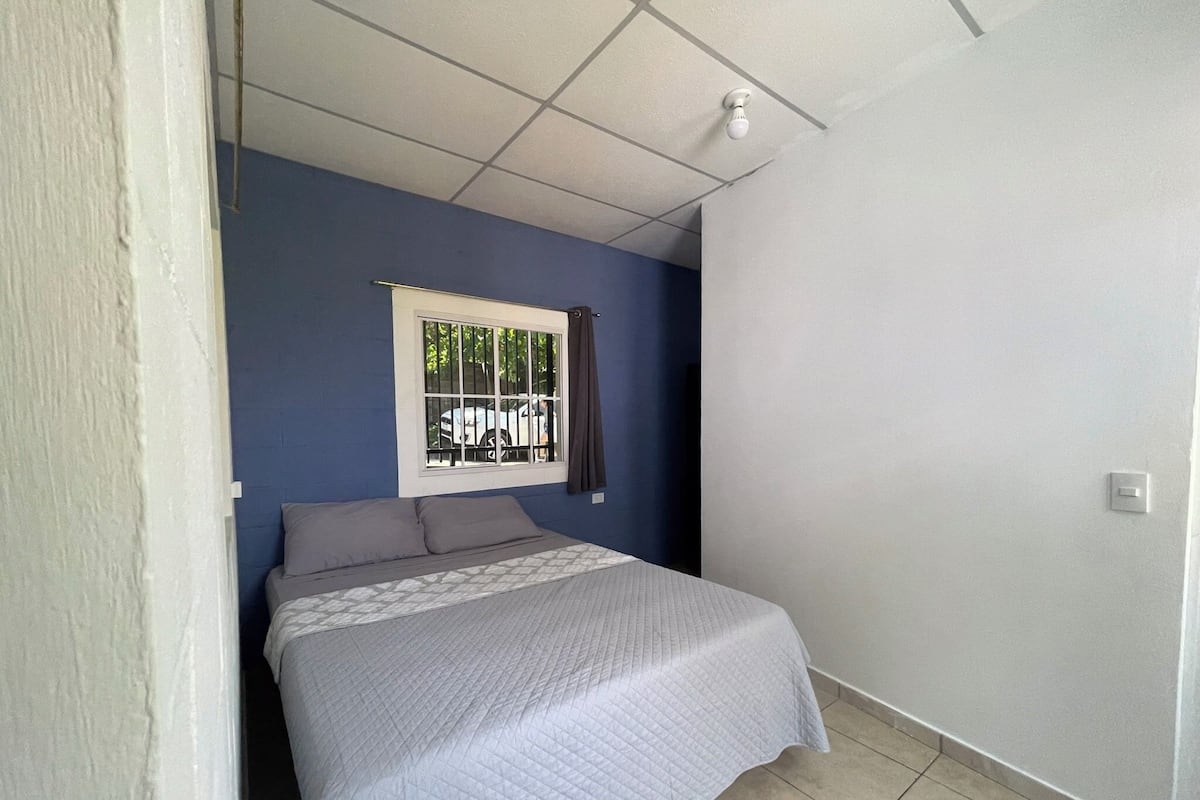 Quarto com cama, janela com grades, luminária no teto e interruptor de parede.