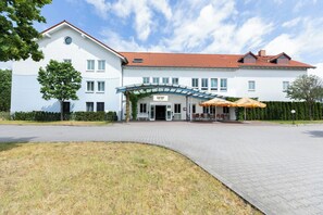 Hotel com fachada branca, telhado vermelho e entrada coberta com área de estar externa.