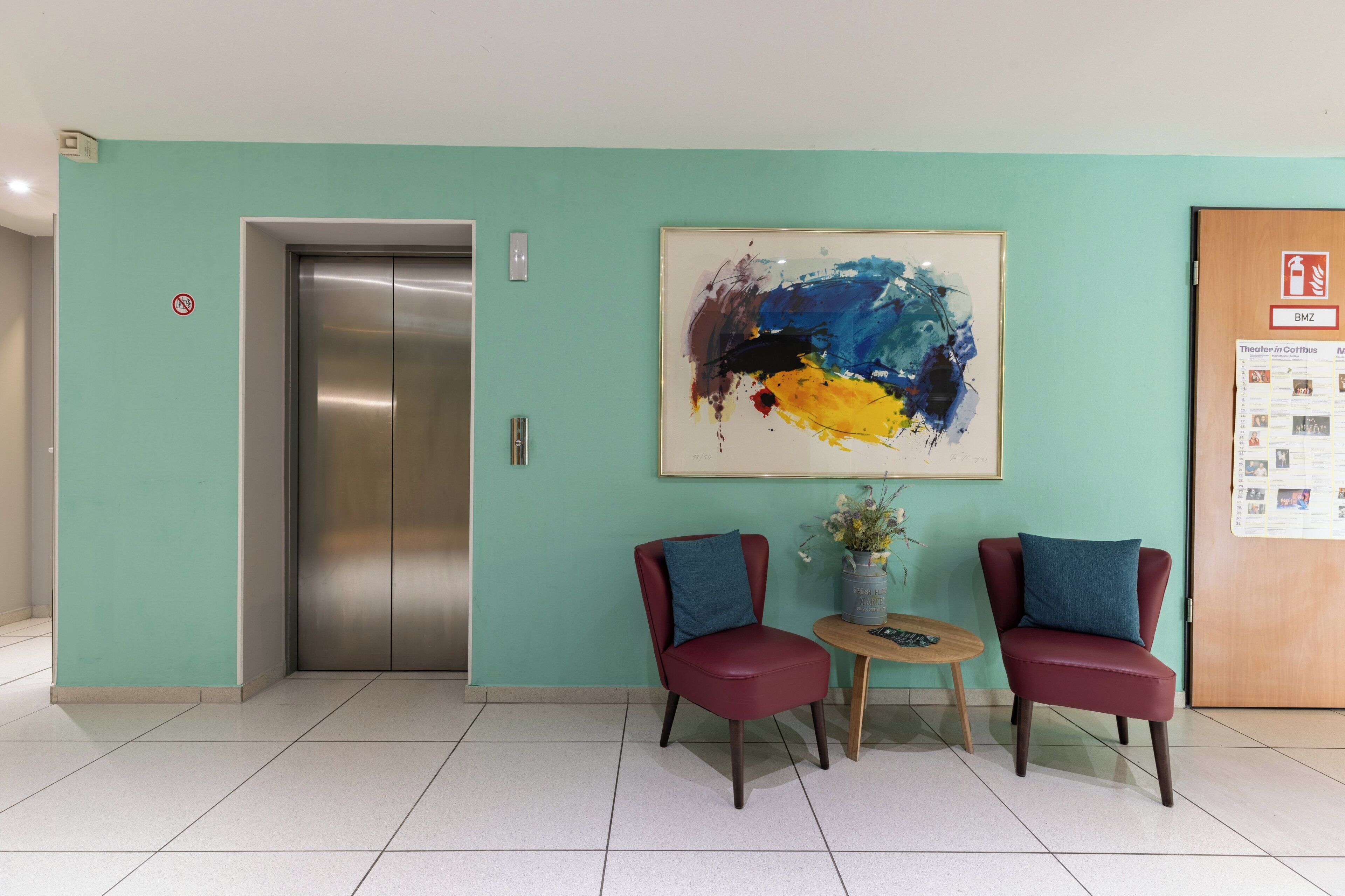 Eine Lobby mit Aufzug, zwei Sessel, einem Tisch mit einer Vase und einem an der Wand befestigten abstrakten Gemälde.