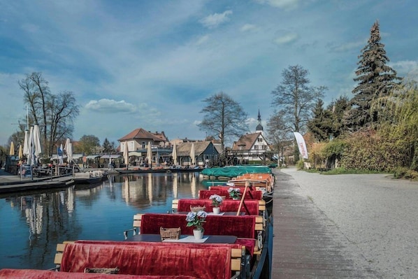 A riverside restaurant with tables and chairs, a dock with boats, and buildings in the background.