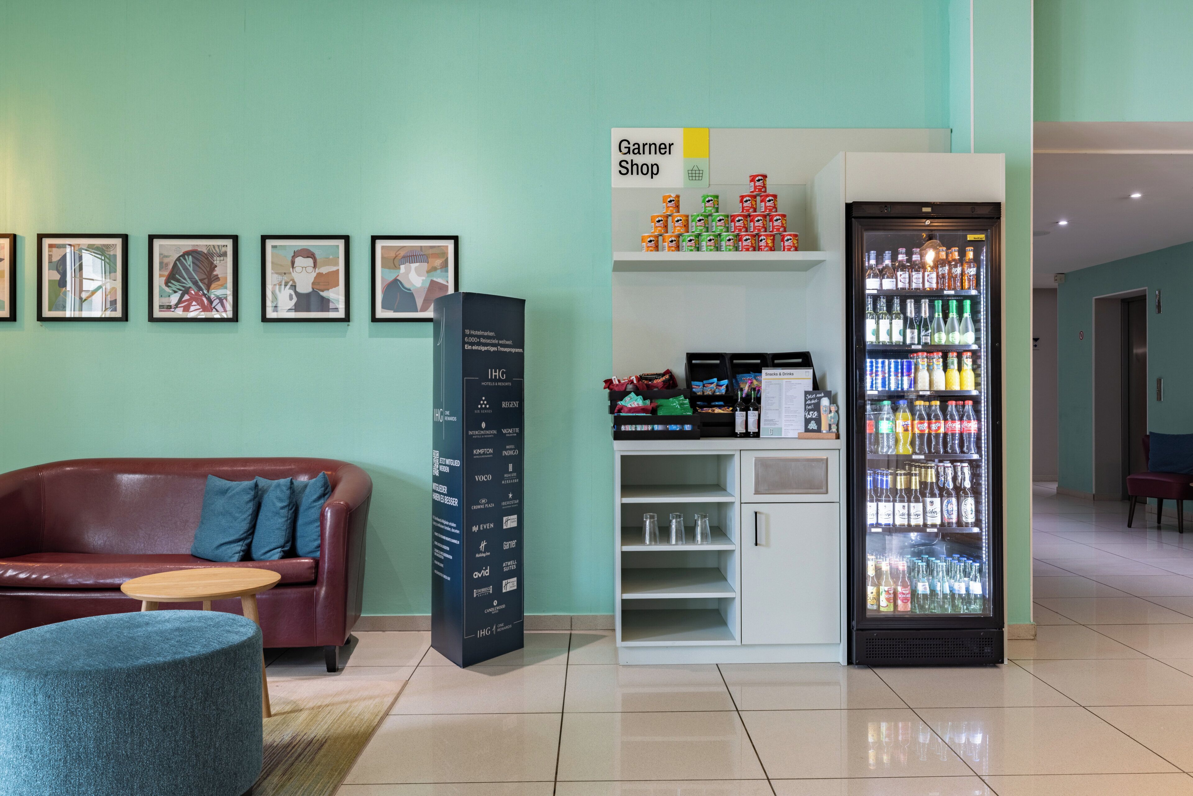 Eine Hotelhalle mit einem roten Sofa, einem kleinen runden Tisch, einem mit Getränken gefüllten Kühlschrank, einem Garner Shop Schild und gerahmten Kunstwerken an der Wand.