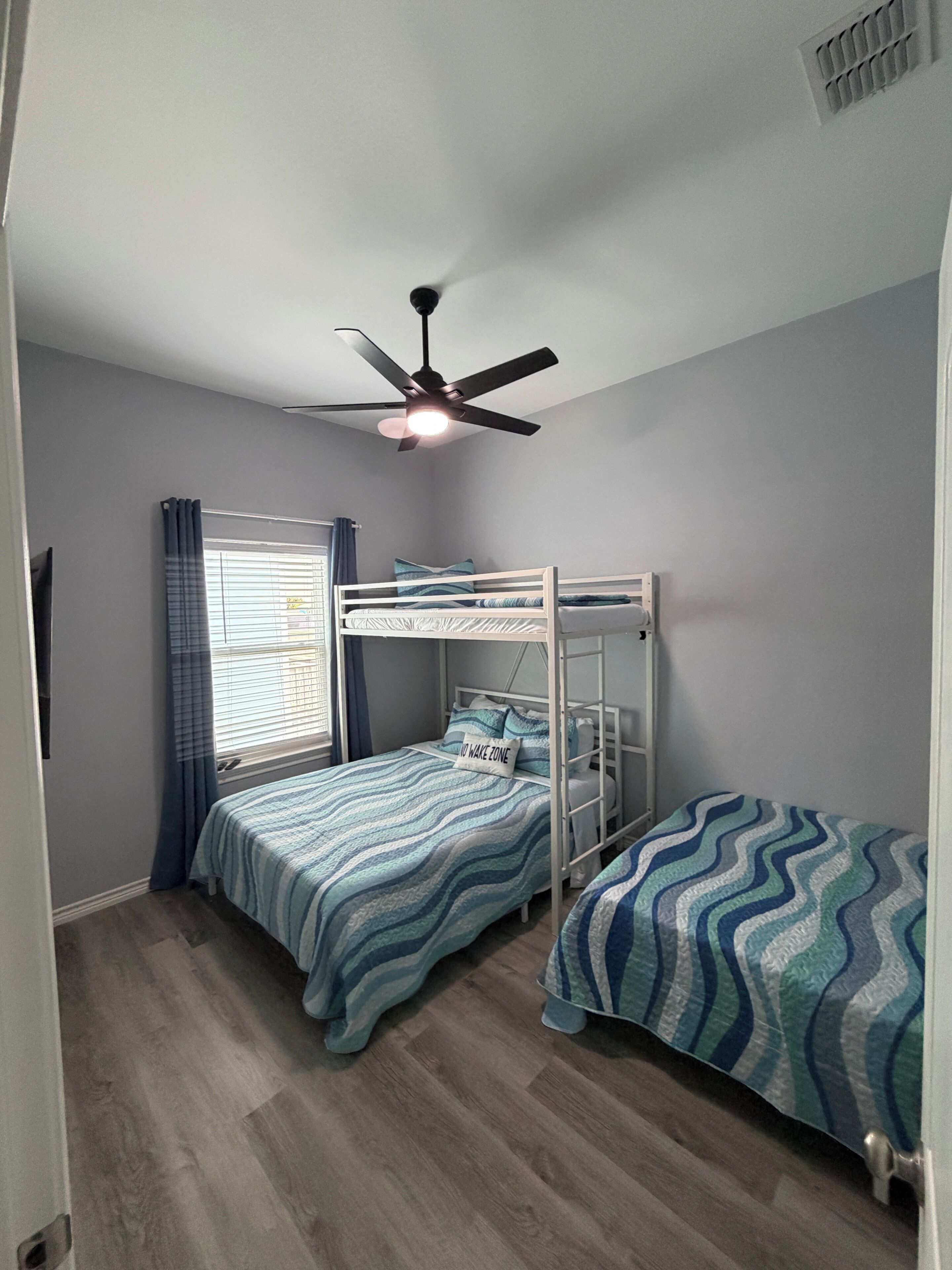 Une chambre avec un lit superposé, un ventilateur de plafond, une fenêtre avec des stores et une porte.