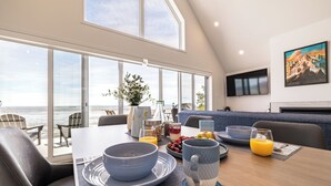 Un comedor con vista al océano, un sofá, un televisor y alimentos para el desayuno sobre la mesa.