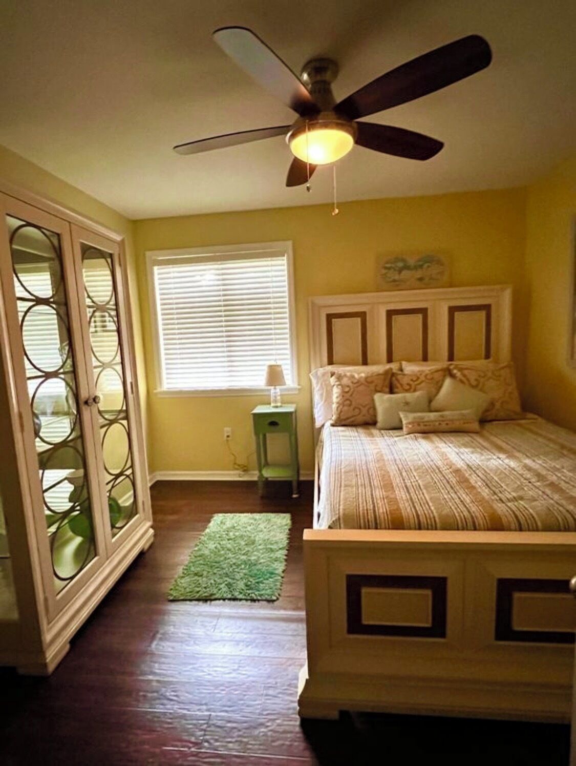 A bedroom with a bed, a ceiling fan, a window with blinds, a green rug, and a wooden floor.