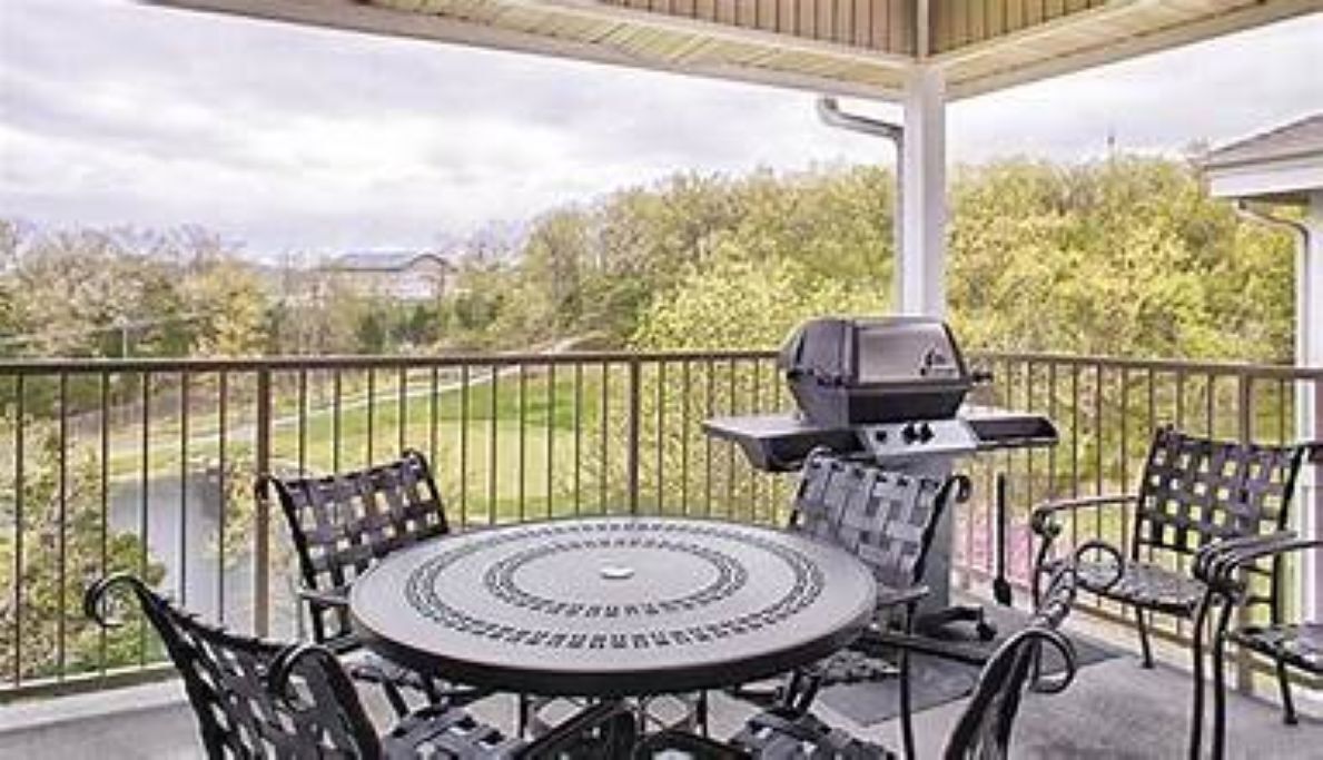 A covered patio with a round table and chairs, a grill, and a view of a green landscape.