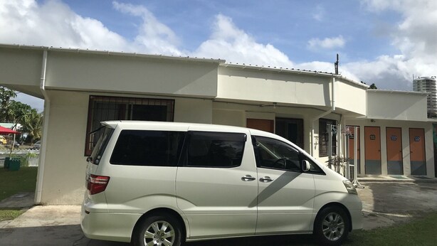 A white van parked in front of a building with a flat roof and a large water tank on the roof. - The Barre (Koror)