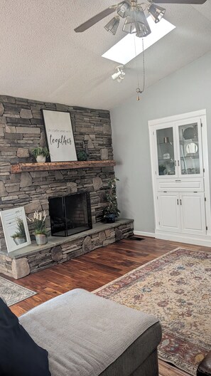 A living room with a stone fireplace, a ceiling fan, a mounted TV, and a patterned rug. - Cozy 2-bedroom house in delightful Marion with AC (Marion)