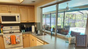 Una cocina con microondas, estufa y lavamanos, un ventanal con vistas a mobiliario exterior, y un cartel con las normas de la casa.