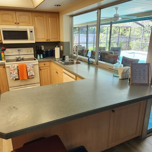 A kitchen with a microwave, stove, and sink, a large window with a view of outdoor furniture, and a sign with house rules. - Poolside Paradise (Safety Harbor)