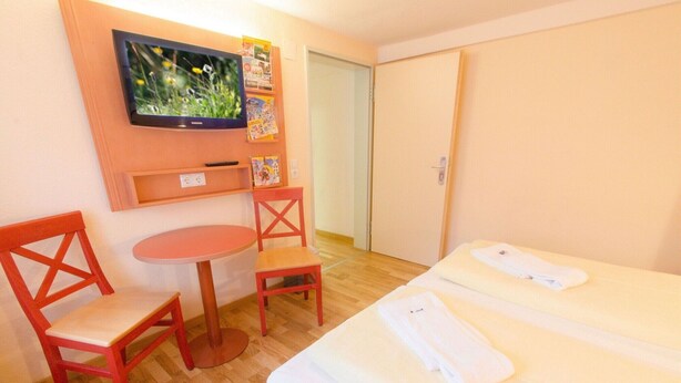 A hotel room with a bed, a TV mounted on the wall, a small table, two chairs, and a door. - Jufa Meersburg am Bodensee (Meersburg)
