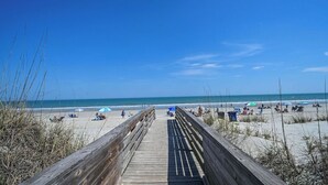 Un paseo marítimo de madera que conduce a una playa con gente y sombrillas.