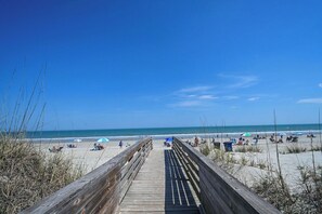 Ein hölzerner Steg führt zu einem Strand mit Menschen und Sonnenschirmen.