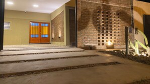 Een moderne gevel met een betonnen pad dat leidt naar een houten deur, een bakstenen muur met een decoratief patroon, en een cactuss. 