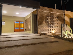 A modern building entrance with a concrete pathway leading to a wooden door, a brick wall with a patterned design, and a cactus plant.