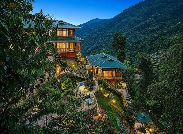 Uma pousada de dois andares com um caminho de pedra que leva à entrada, cercada por vegetação exuberante e um cenário montanhoso.
