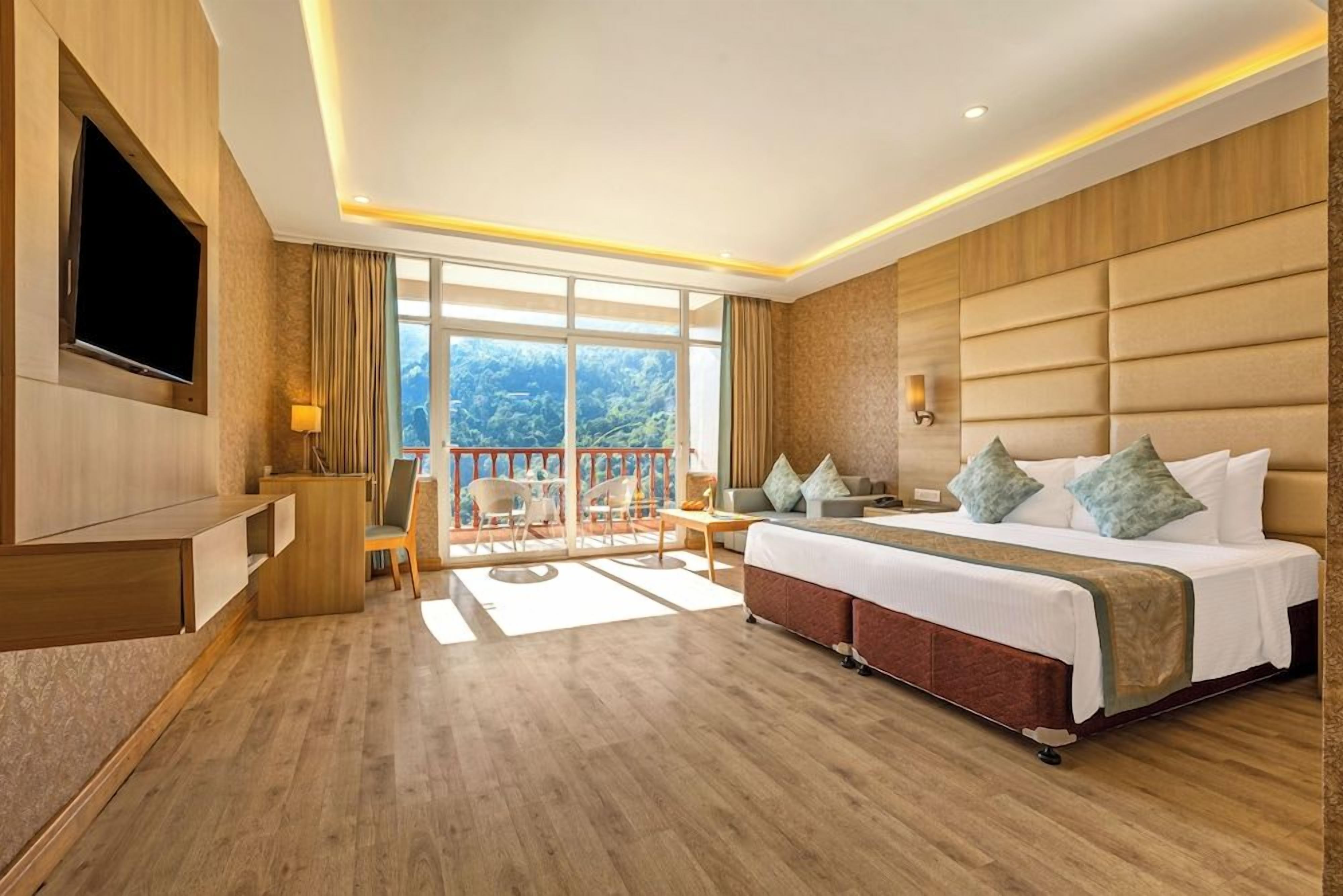A hotel room with a large bed, a TV, a balcony with a view, and a wooden floor.