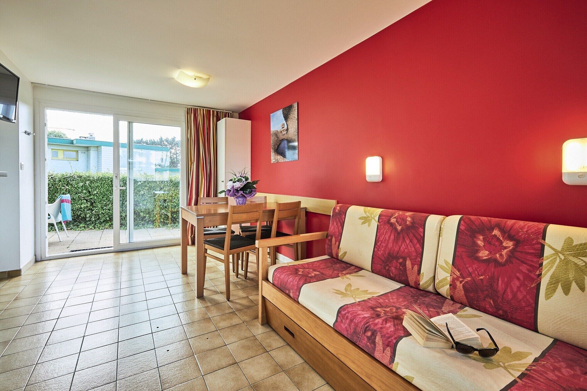 Ein Zimmer mit roter Wand, einem Holzsofa, einem Esstisch mit Stühlen, einer Glastür zum Außenbereich und einem kleinen Topfpflanzen.