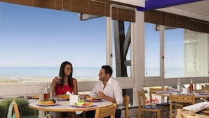 Un espace repas offrant une vue sur la plage, avec des tables rondes, des chaises et un buffet proposant des plats et des boissons.