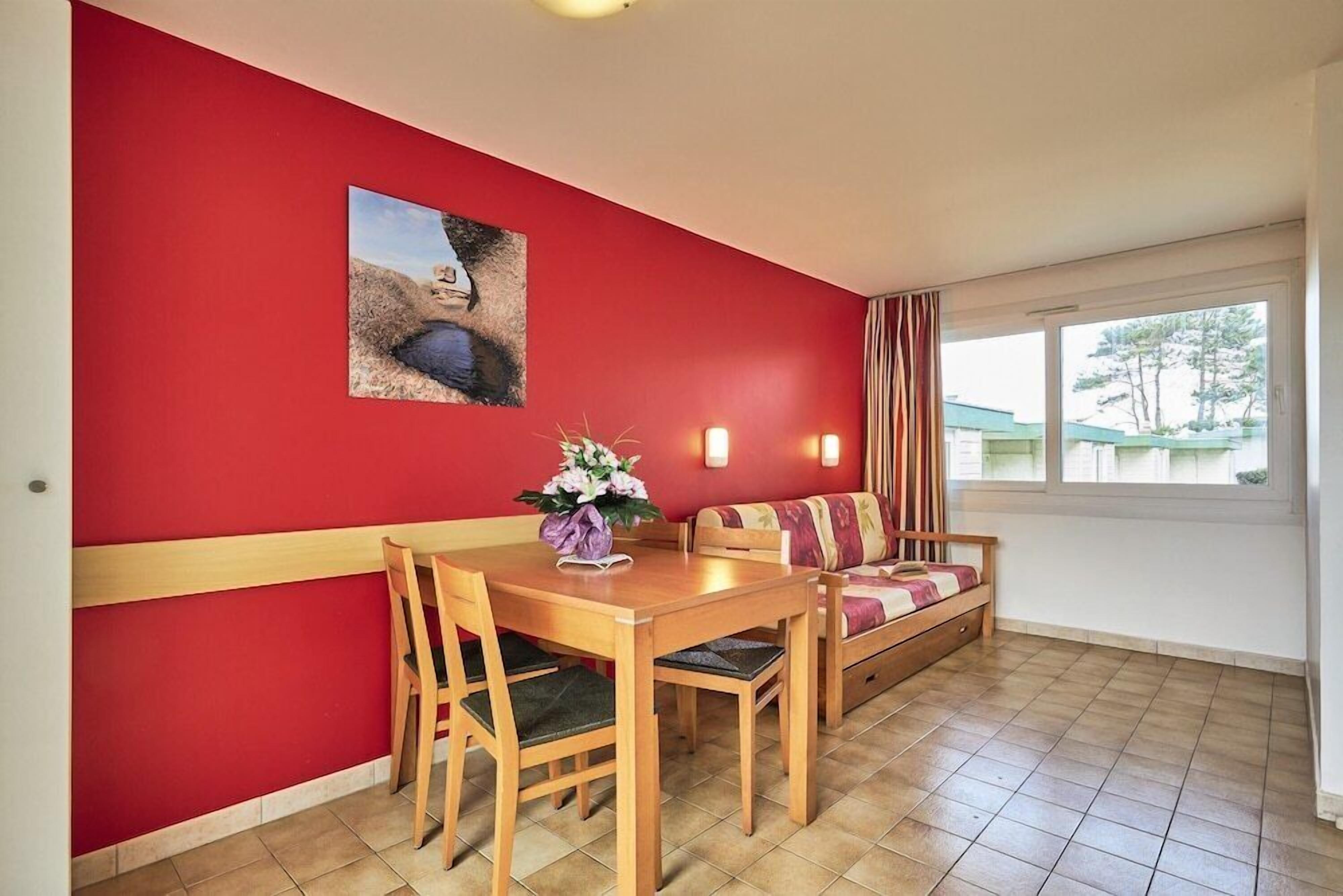 A dining area with a red wall, a wooden table, chairs, a sofa, and a window with curtains.