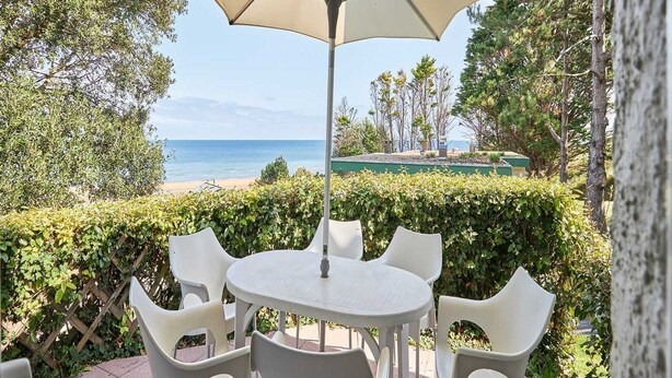 Une terrasse avec une table et des chaises, un grand parasol et une vue sur la plage et l’océan.