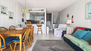 Ein Essbereich mit Holztisch und -stühlen, im Hintergrund eine Küche, eine Couch mit gemusterten Kissen und ein Teppich mit Muster.