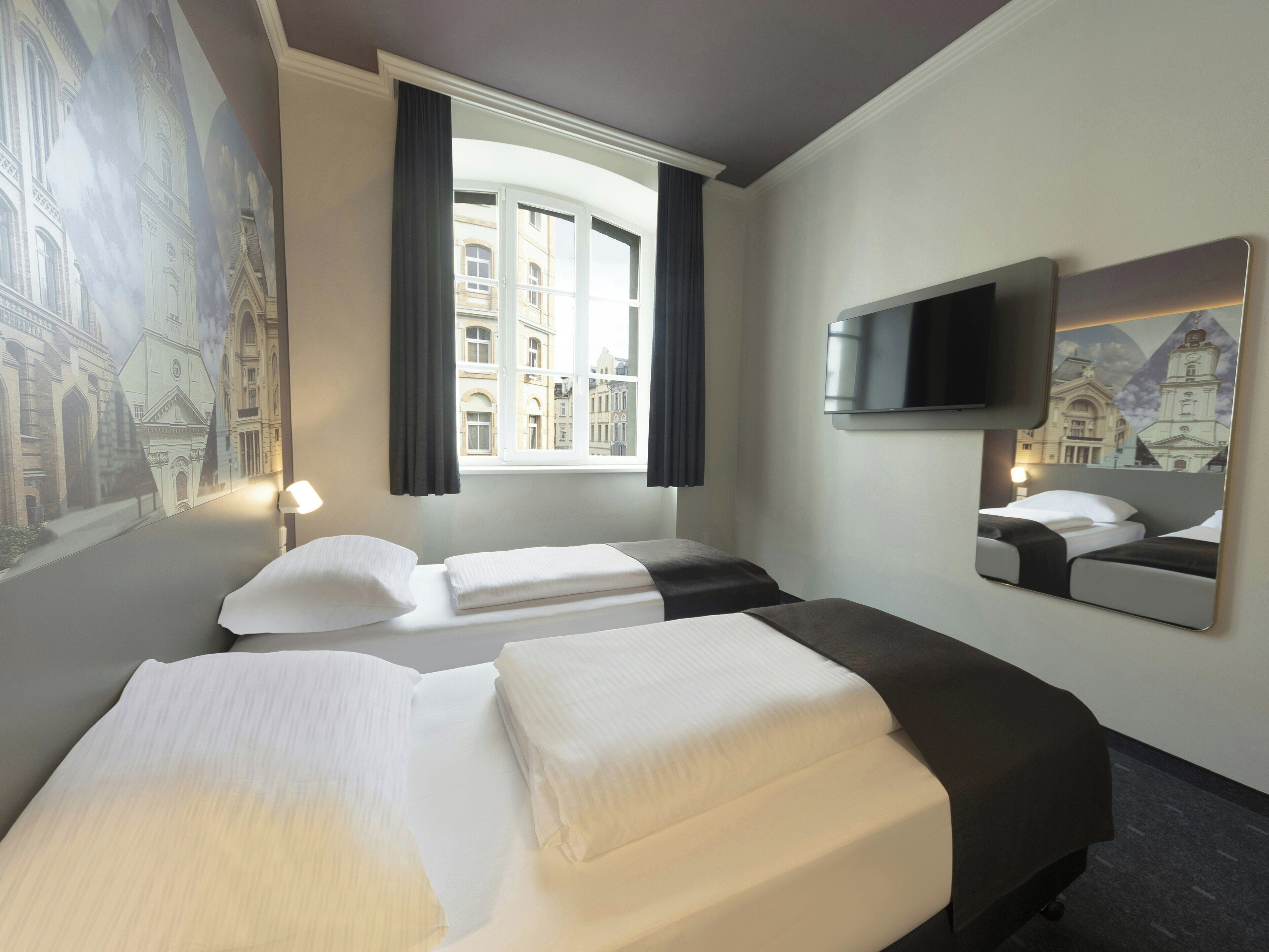 Une chambre d’hôtel avec deux lits, une grande fenêtre, un téléviseur à écran plat et une fresque représentant un bâtiment.