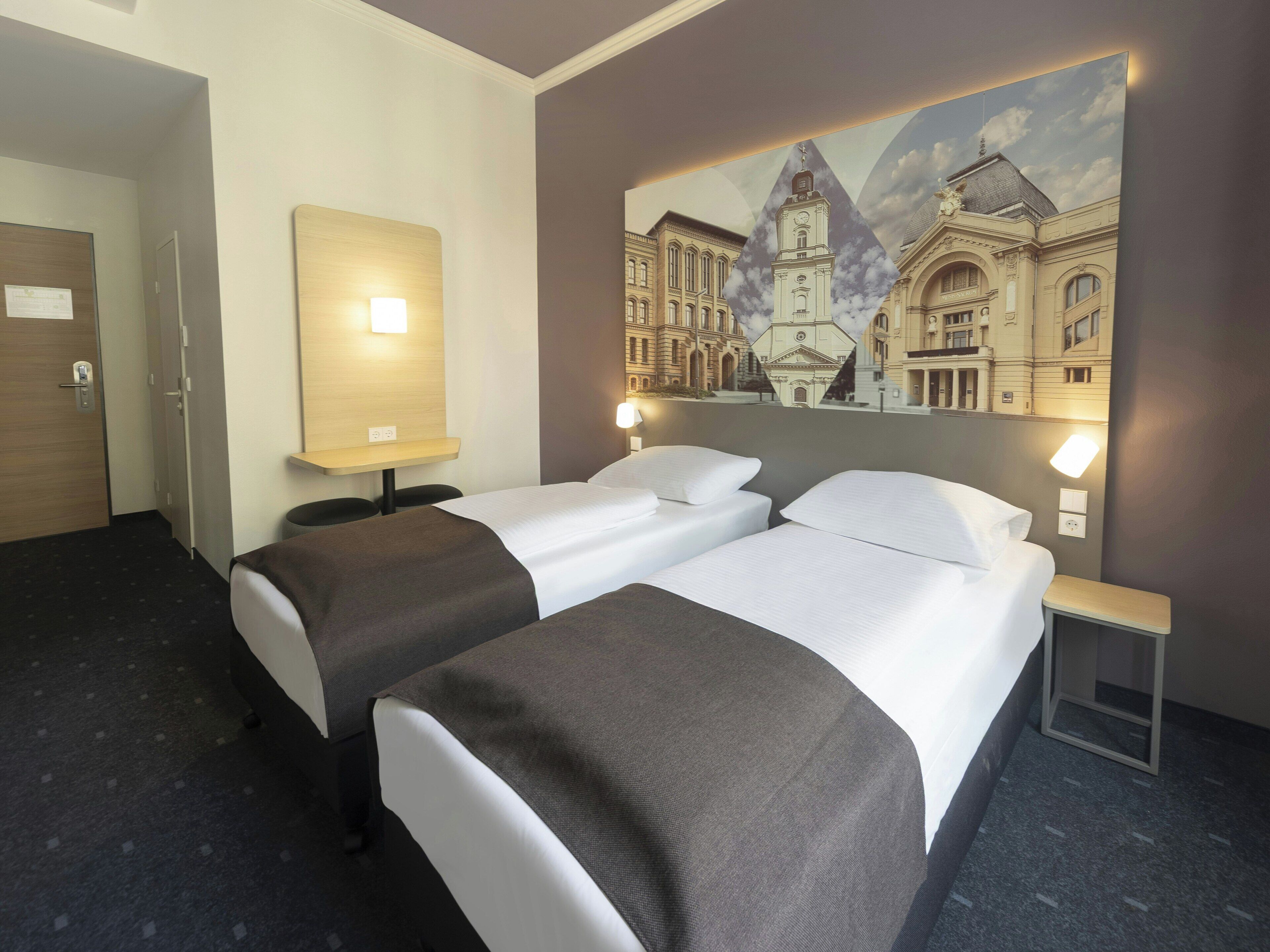 Una habitación de hotel con dos camas, una mesita de noche, una lámpara de pared y un cuadro de un edificio.