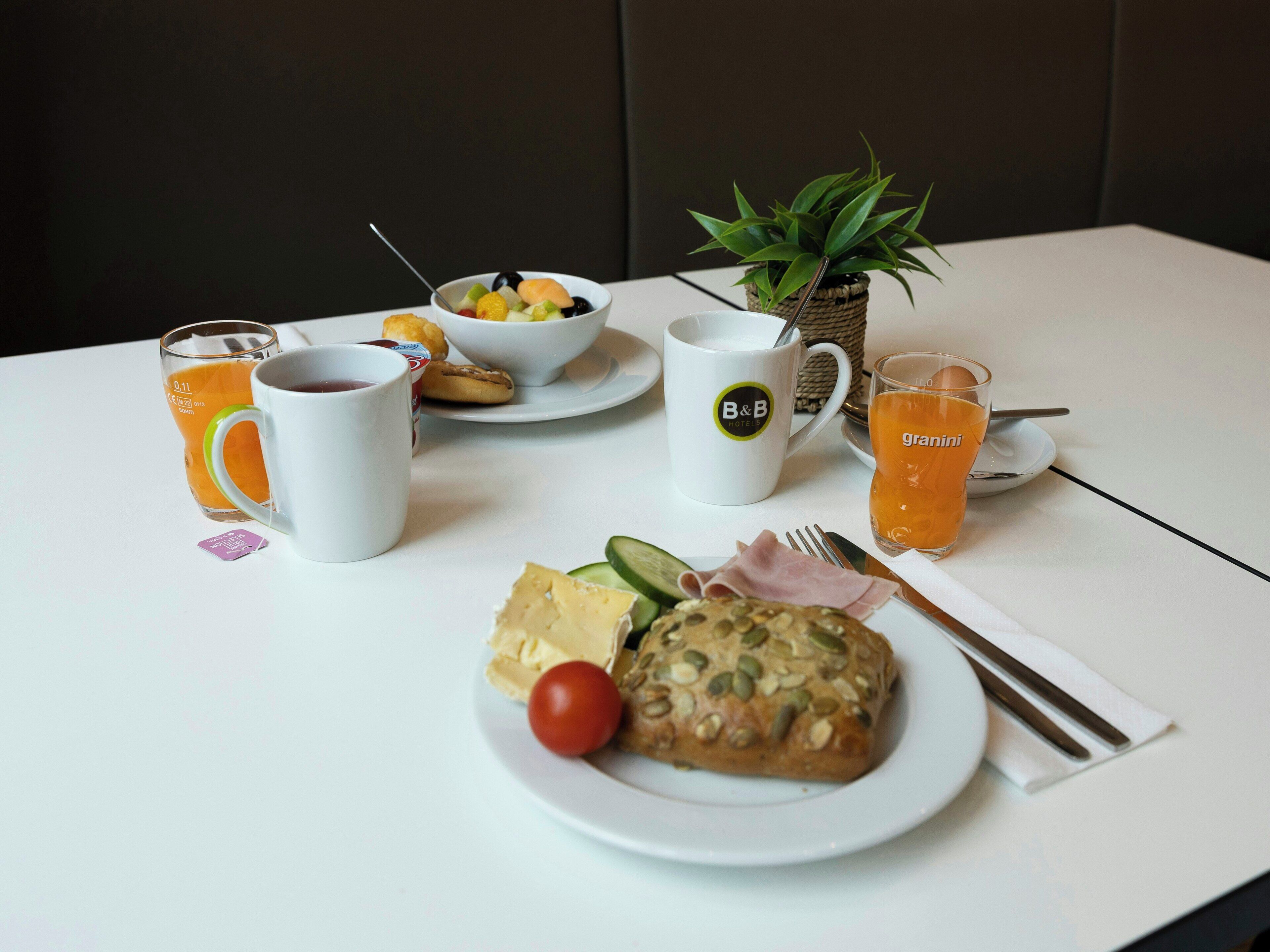 Una mesa con alimentos para el desayuno, incluyendo un plato de comida, un tazón de fruta, un vaso de jugo de naranja, una taza de café y una mug con el logo del hotel.