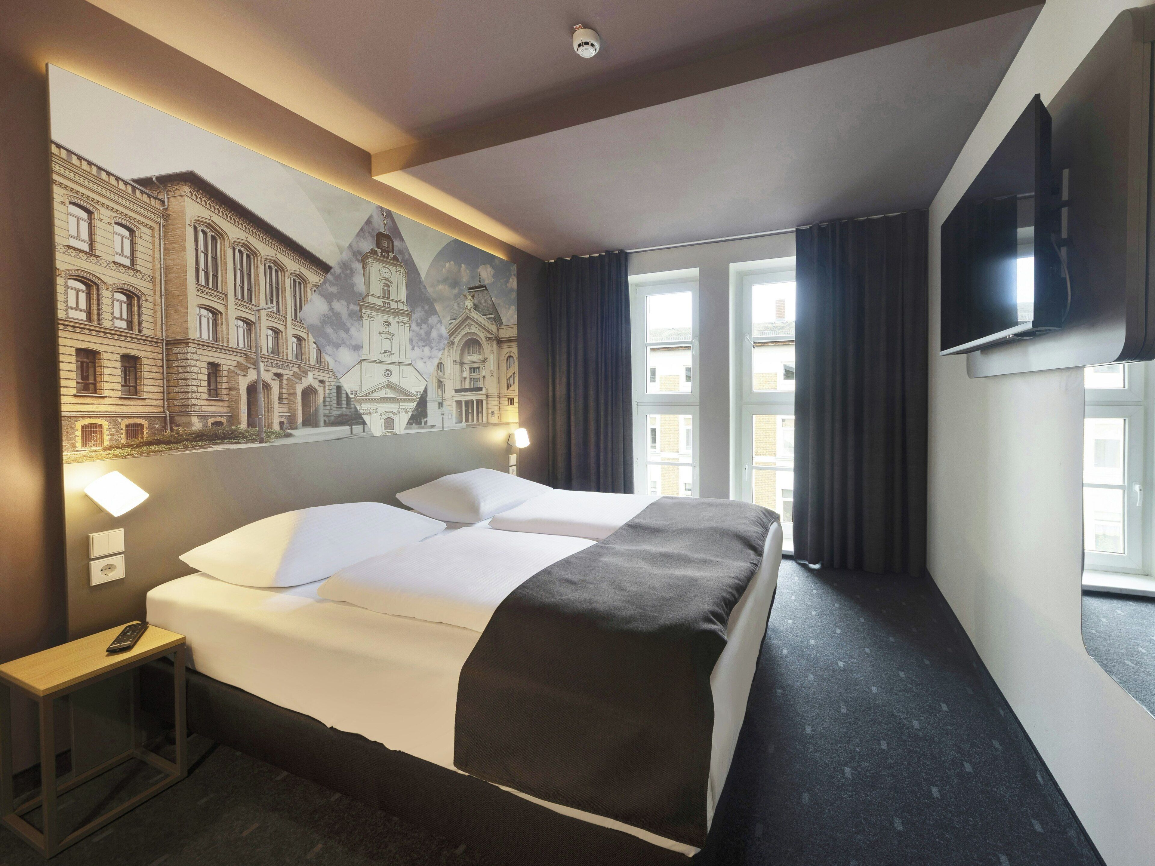A hotel room with a large bed, a flat-screen TV, a window with curtains, and a wall-mounted lamp.