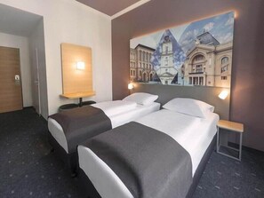A hotel room with two beds, a painting of a building on the wall, and a nightstand with a lamp. - The Royal Inn Regent (Gera)