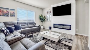 A living room with a grey sectional sofa, a white coffee table, a mounted TV, and a fireplace. - The Hills in Lorson Ranch (Colorado Springs)