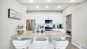A modern kitchen with white cabinets, a granite countertop, stainless steel appliances, and a bar area with white stools. - The Hills in Lorson Ranch (Colorado Springs)
